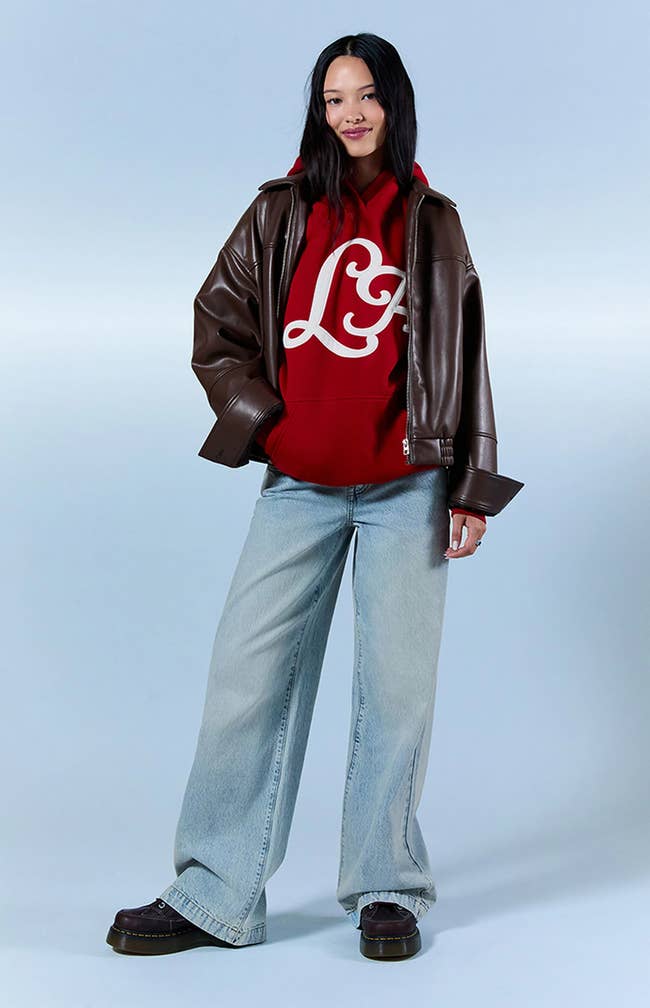 Person modeling wide-leg jeans, a reddish  hoodie with a ample  logo, and an oversized leather jacket, suitable for a casual buying  outfit