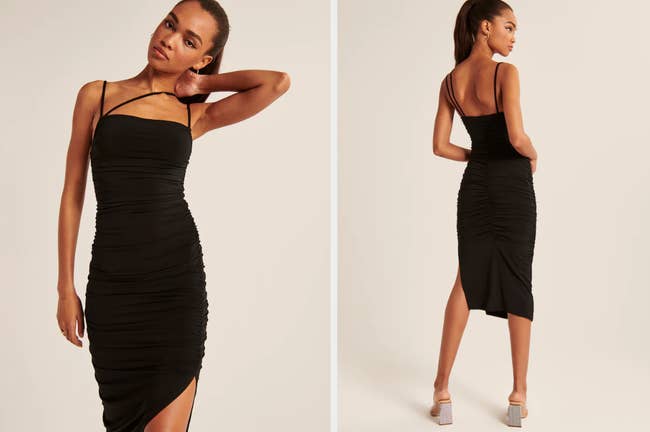 Model wearing black sleeveless bodycon dress with asymmetrical strap across chest, ruched material, and small side slit, model showing back view of product with black heels on a tan background