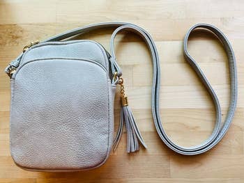 客製化系統 reviewer's light grey crossbody bag with a zippered compartment and tassel detail on the side, shown on a wooden surface