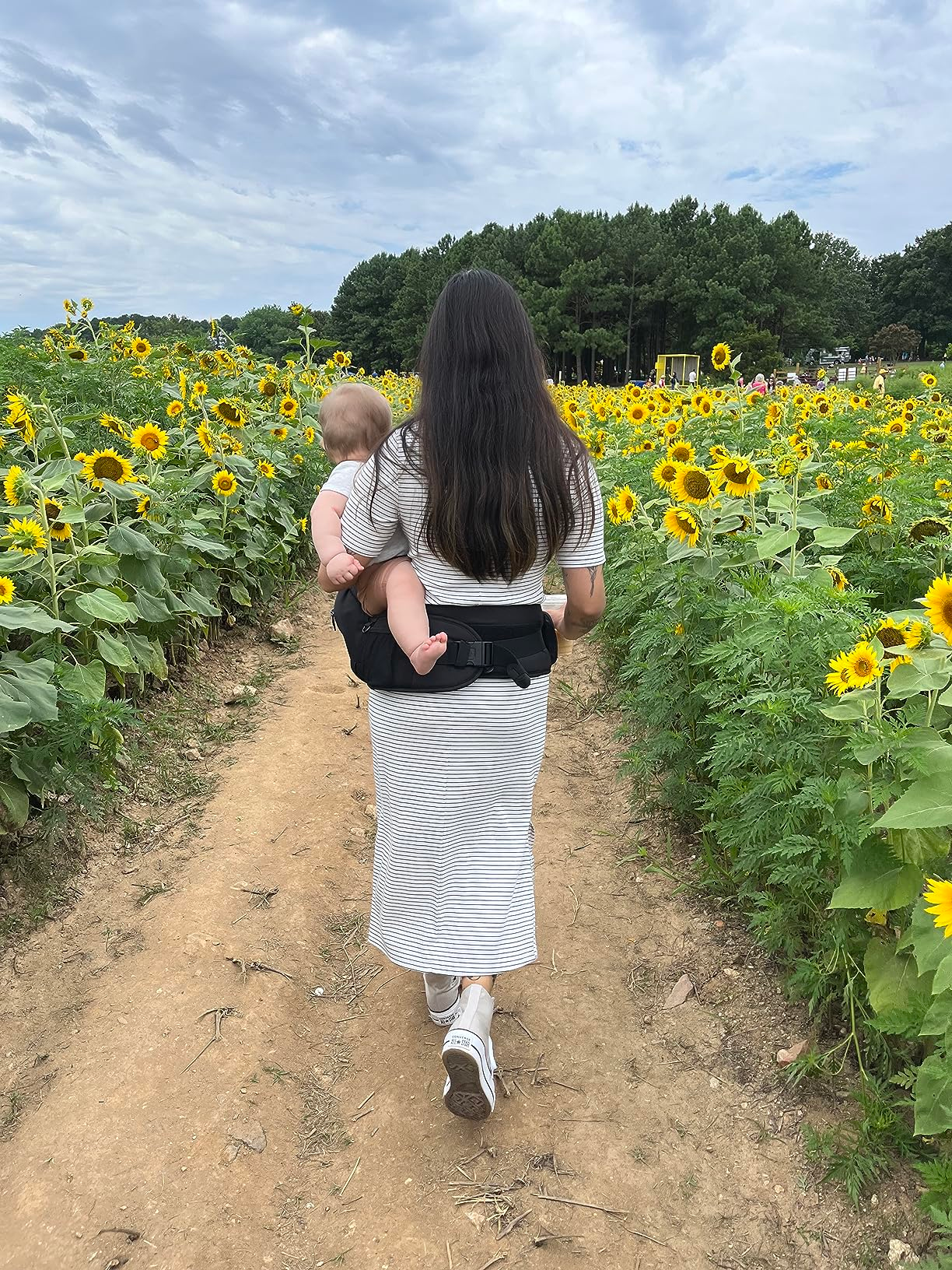Person in a striped dress walks through a sunflower field, carrying a baby in a black carrier on a dirt path