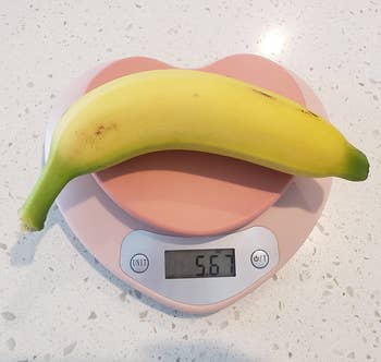 A banana rests on a digital kitchen scale displaying 56.7 grams. The scale is on a solid surface