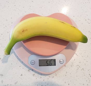 A banana rests on a digital kitchen scale displaying 56.7 grams. The scale is on a solid surface