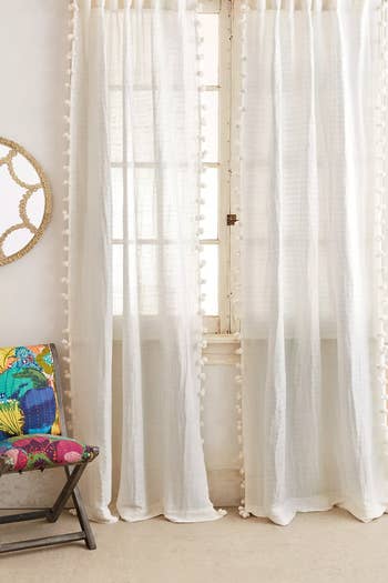 White sheer curtains with pom-pom details, beside a chair with a colorful cushion