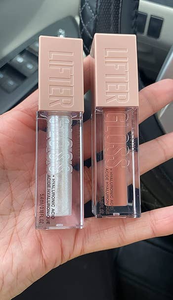 reviewer photo of them holding two tubes of the lip gloss, one in a clear shade and the other in a nude-brown