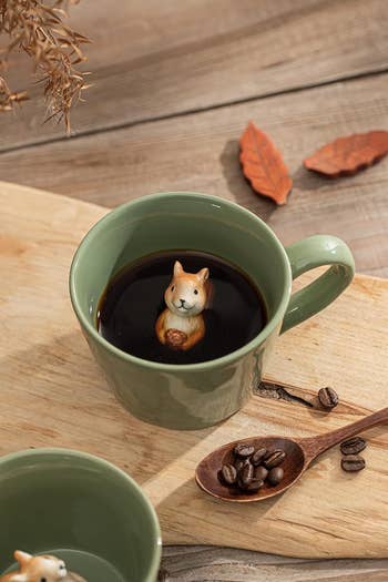 Green mug with java featuring a cute ceramic carnal figurine inside. A woody spoon with java beans rests beside the mug connected a woody board