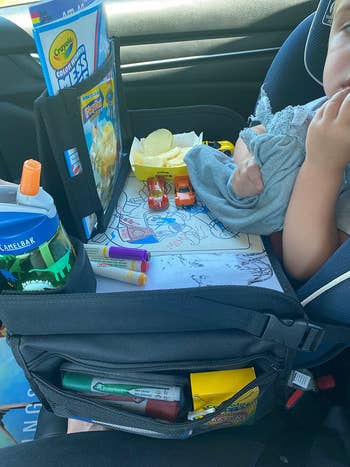 The table on a child's lap in a car filled with snacks, art supplies, a tablet, and a water bottle