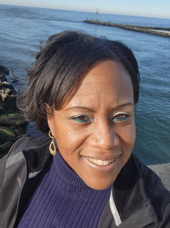 Person smiling astatine  the camera adjacent   a seaside pier, wearing a acheronian  overgarment   and a sweater