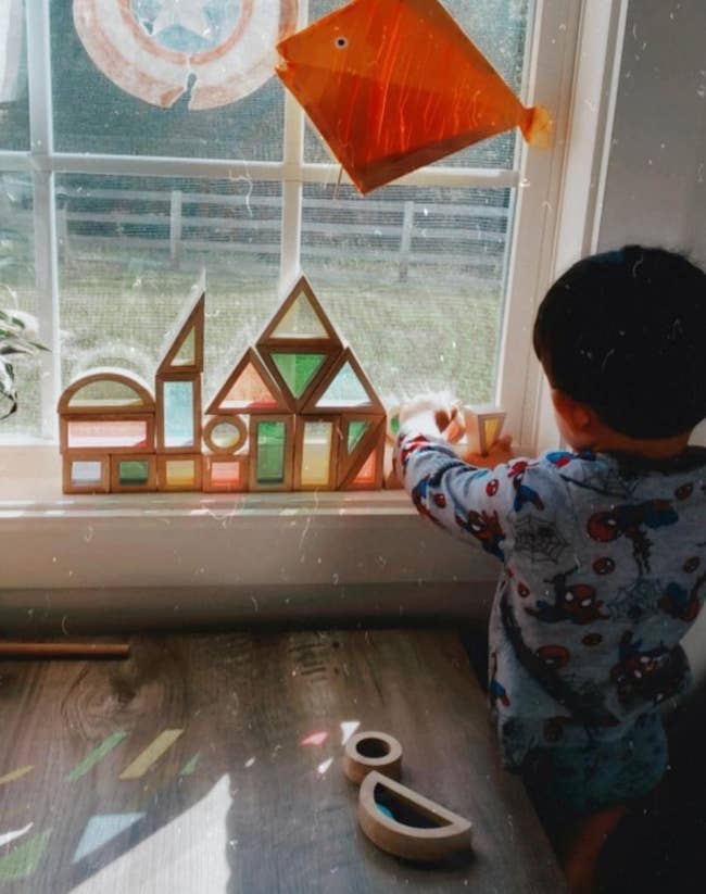 Child plays with geometric shapes near window with a kite decoration, suggesting creative play-focused products