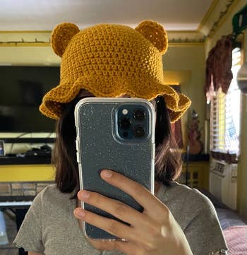 Model wearing yellow teddy-shaped crochet bucket hat holding a smartphone