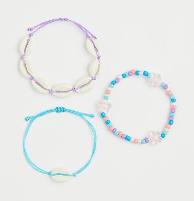 Purple string bracelet with white shells, blue string bracelet with single shell, blue, purple, pink, and white beaded bracelet with white daisy beads on a white background