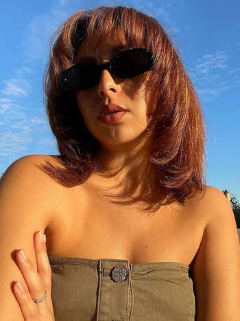 A reviewer with medium-length hair, wearing sunglasses, a strapless top with a decorative button, and a ring poses outdoors on a sunny day