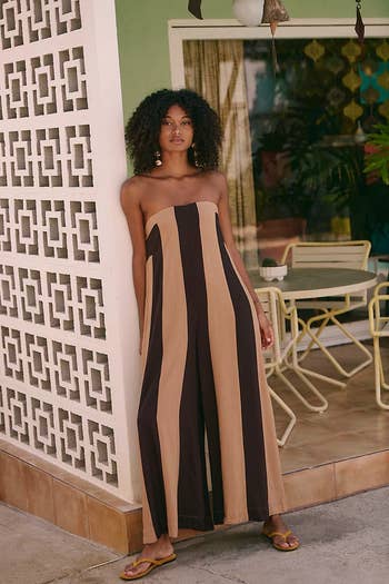 Person in a strapless, striped jumpsuit and sandals leans against a patterned wall near outdoor seating area