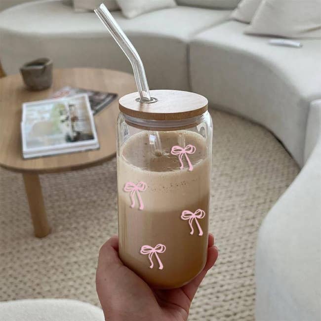 Hand holding a jar with a wooden lid and straw, decorated with bow illustrations, filled with a coffee beverage. Modern living room in the background