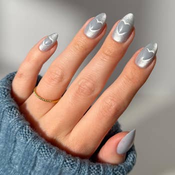 Person's manus  with long, almond-shaped nails featuring a metallic decorativeness  and bosom  design. The manus  wears a bluish  sweater and a bladed  golden  ring