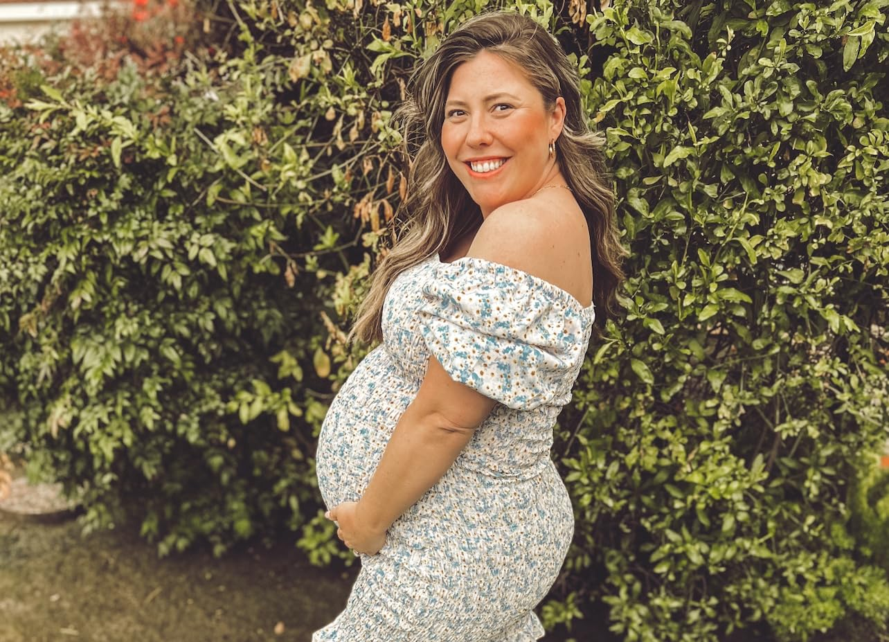 Person smiling outdoors, wearing an off-shoulder floral dress, holding their pregnant belly