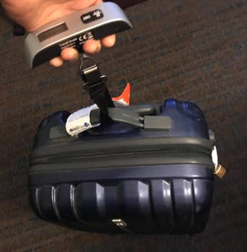 Hand holding a luggage scale with a small, black, wheeled suitcase attached for weighing