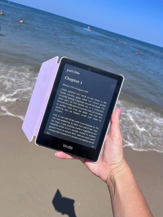 Hand holding a Kindle against a beach backdrop with the ocean and swimmers visible, displaying an opened e-book