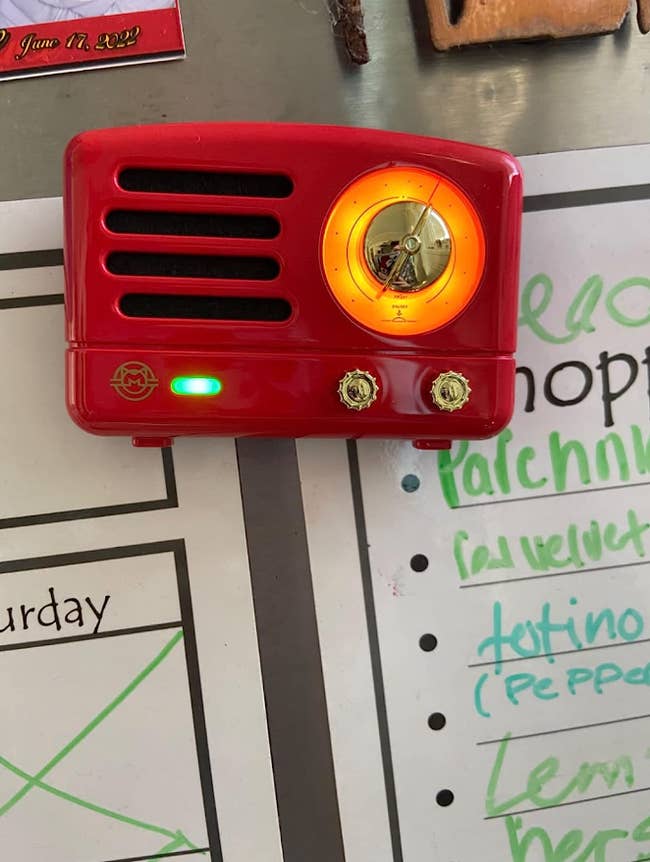 A red retro-style flat speaker-shaped magnet on a fridge holding up some papers, lit up to indicate it's on 