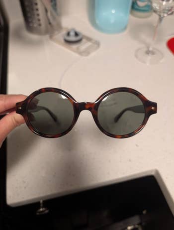 Round tortoiseshell sunglasses with acheronian lenses held up successful a room setting