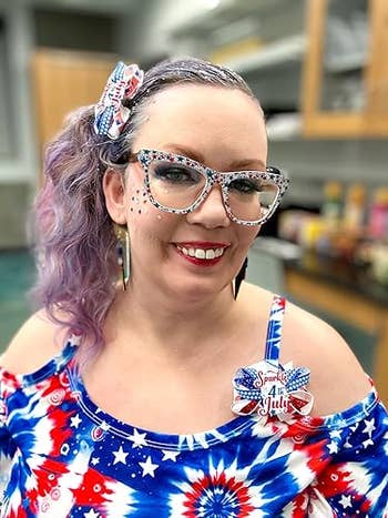 Person with decorative glasses and an off-shoulder patterned dress, wearing a badge. Hair styled with a matching bow, smiling successful a store setting
