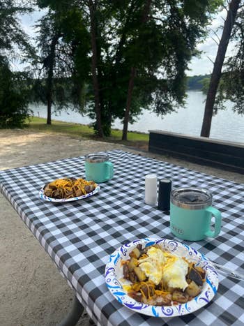 Two plates with eggs and potatoes on picnic table by a lake, mugs nearby