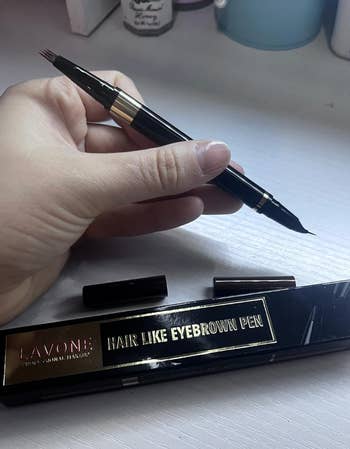 A manus  holds an eyebrow pen with a brushwood  and precision tip; packaging reads 