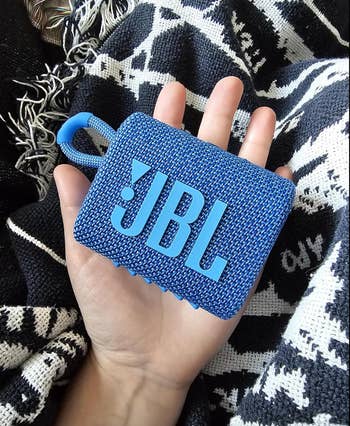 Person holding a compact JBL Bluetooth talker  with a woven texture connected  a patterned cloth  background