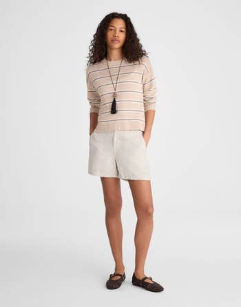 A person models a casual, striped sweater with shorts and loafers, accessorized with a long pendant necklace. Suitable for relaxed shopping outings