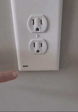 A person's finger is pointing towards a smart electrical outlet with a built-in LED indicator light