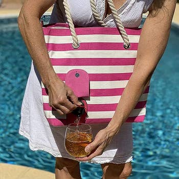 Person in a sleeveless white dress by a pool, holding a pink and white striped tote bag with rope handles, pouring a drink from a spout into a glass