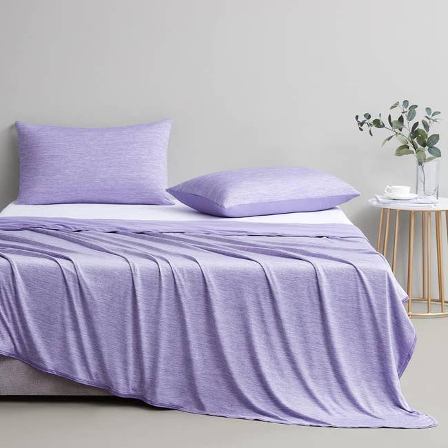 Purple bed set with smooth sheets and pillowcases on a made bed, next to a side table with a potted plant and a cup