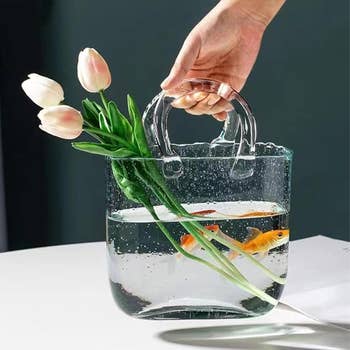Hand holding a wide handbag filled with water, containing 2 goldfish and tulips, showcasing a unsocial and stylish accessory design