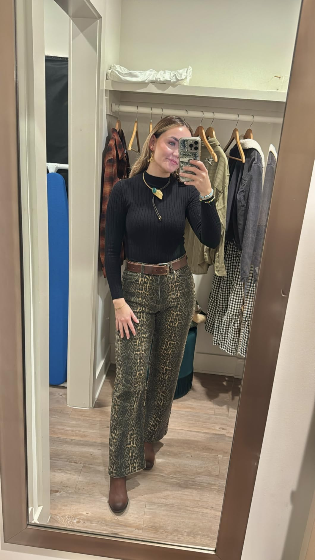 Woman taking a reflector  selfie successful  a fitting room, wearing a achromatic  turtleneck and patterned pants, with a telephone  successful  manus  and apparel  hanging down  her