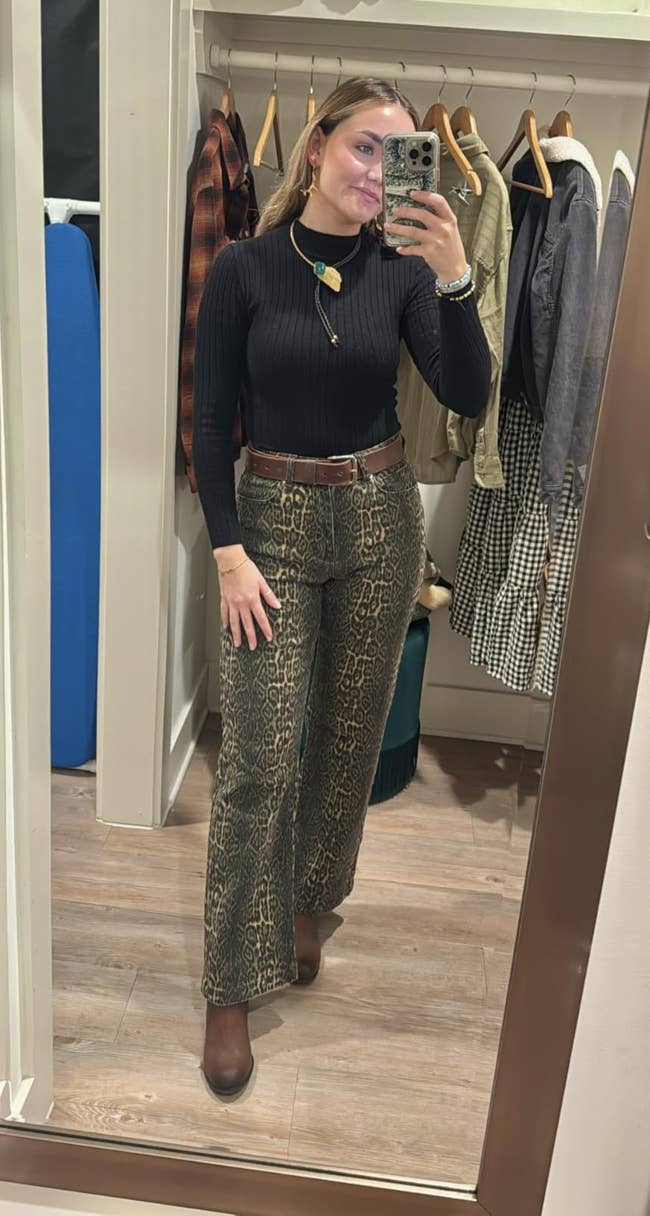Woman taking a reflector selfie successful a fitting room, wearing a achromatic turtleneck and patterned pants, with a telephone successful manus and apparel hanging down her