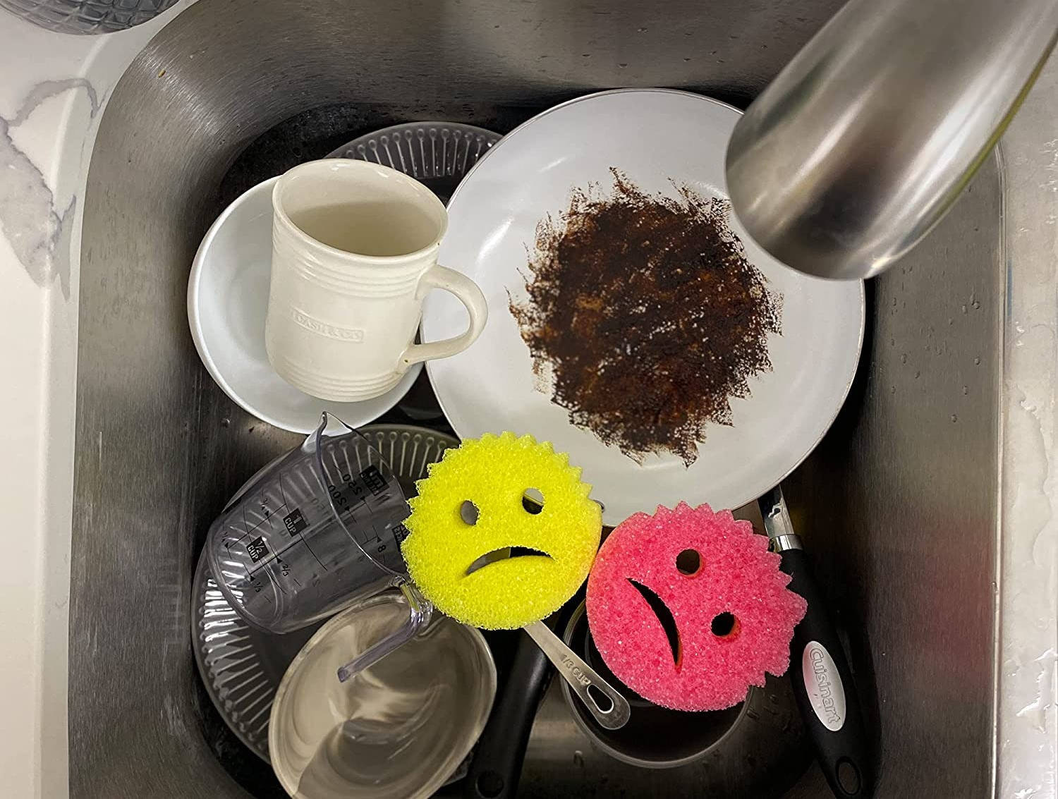 a set of two of face-shaped frowning sponges in a sink of dirty dishes 