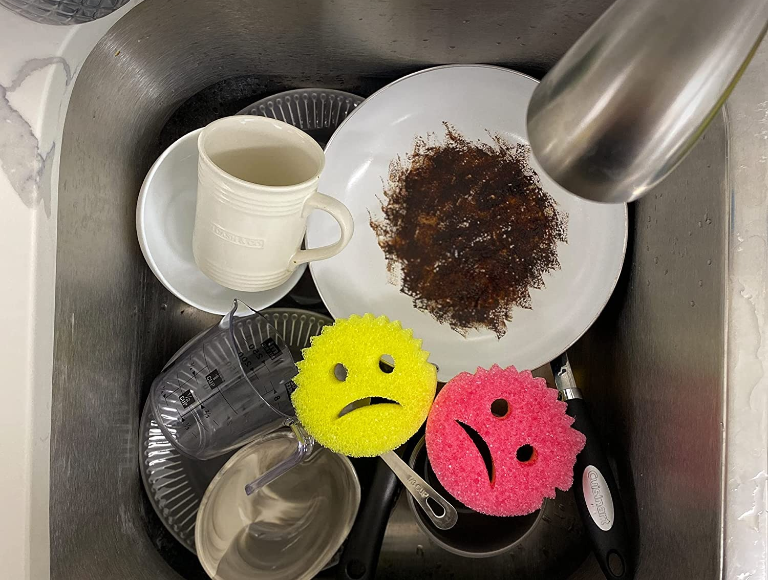 a set of two of face-shaped frowning sponges in a sink of dirty dishes