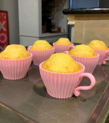 Six freshly baked cupcakes sit inside pink teacup-shaped silicone molds on a baking sheet in a kitchen