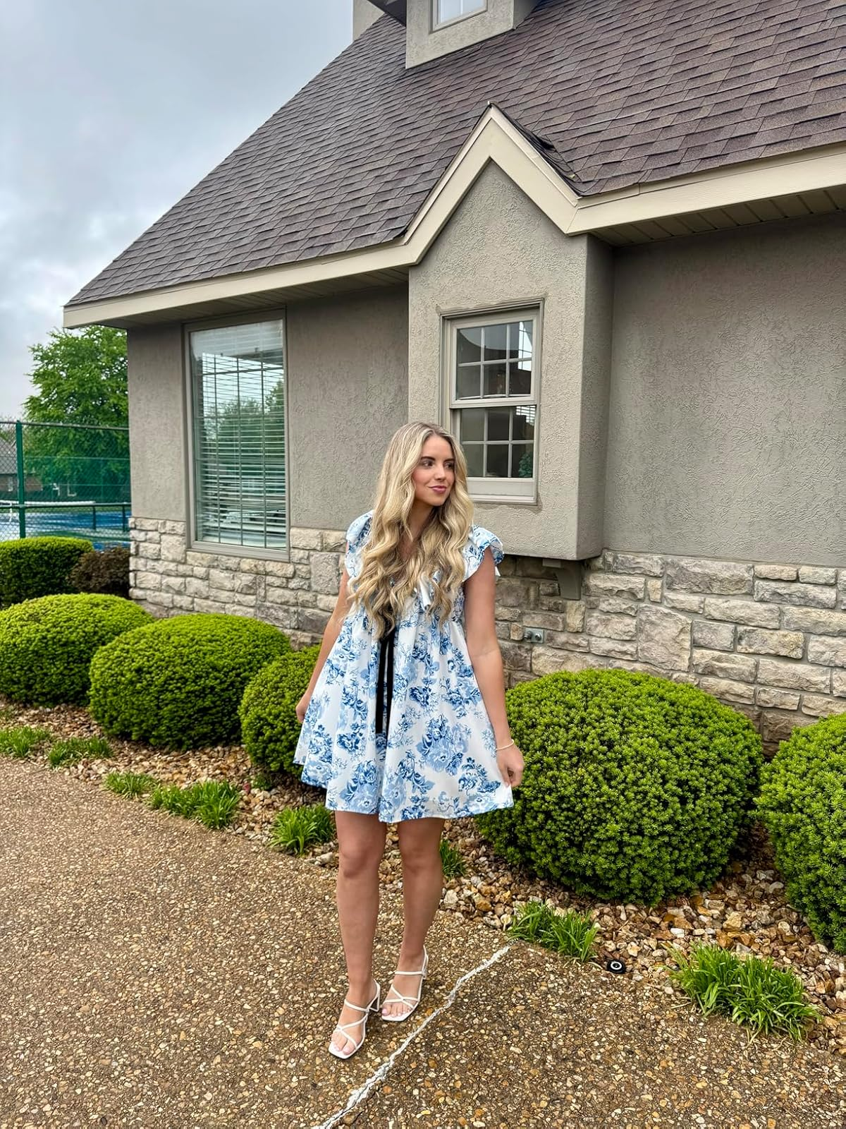Person in a floral dress and heels stands outside a house with shrubbery