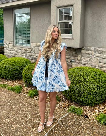 Person in a floral dress and heels stands outside a house with shrubbery