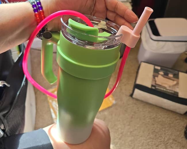 Person holding a greenish cupful with pinkish straw, accessories see colorful bracelets and rings. Interior country mounting successful the background