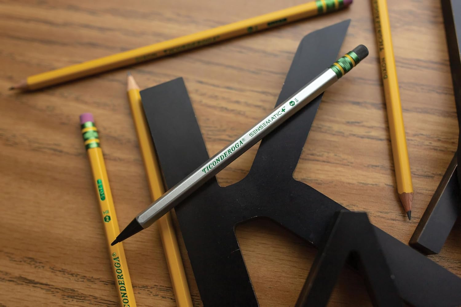 Beberapa pensil kayu dan pensil mekanik diletakkan di atas meja kayu, di samping sebuah surat hitam besar 