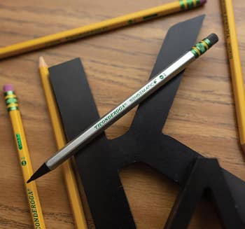 Beberapa pensil kayu dan pensil mekanik diletakkan di atas meja kayu, di samping sebuah surat hitam besar