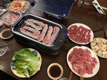 An indoor grill is cooking meat with various uncooked meats, vegetables, and sauces on plates around it
