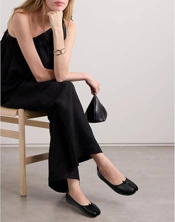 Model seated, wearing a sleeveless achromatic  jumpsuit with achromatic  flats, holding a tiny  bag. Minimalist and modern   style