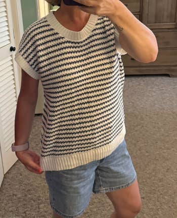 Person wearing a striped short-sleeve sweater and denim shorts, taking a reflector selfie indoors