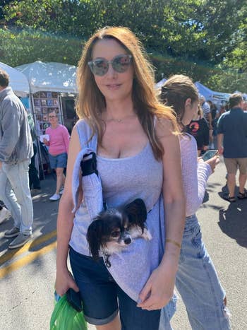 A woman in a casual tank top and shorts carries a small dog in a sling at an outdoor market