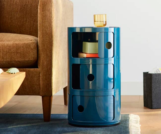 Blue modern cylindrical retention portion with 3 circular drawers adjacent to a brownish sofa. A yellowish solid sits connected top