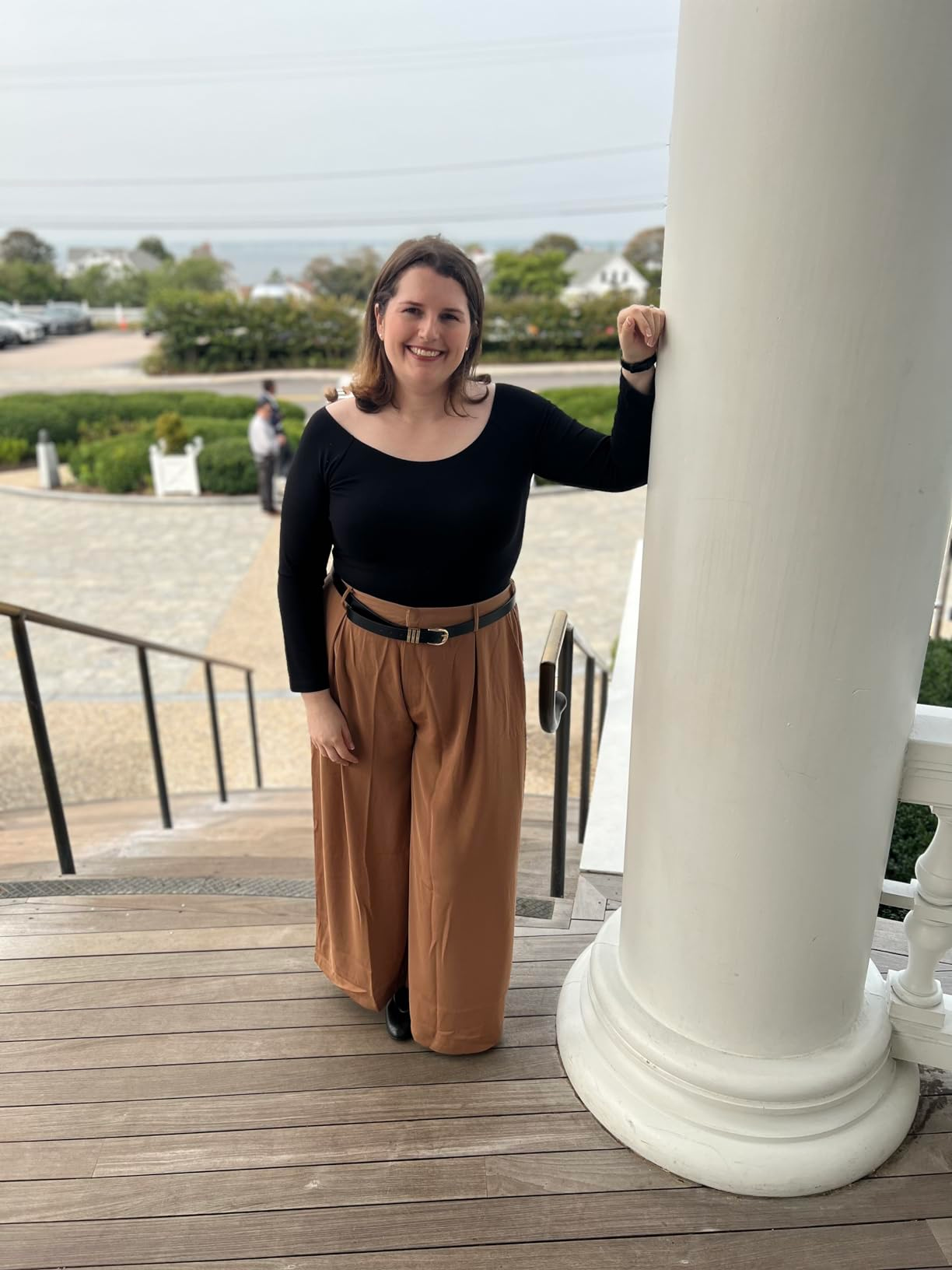 Person stands on steps by a large pillar, wearing a chic black top and wide-legged trousers, smiling casually