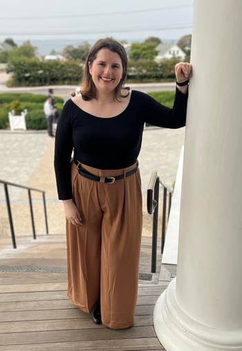 Person stands on steps by a large pillar, wearing a chic black top and wide-legged trousers, smiling casually