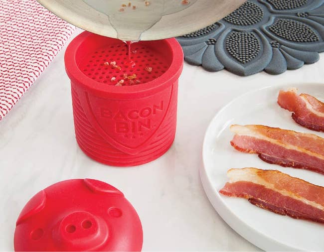 bacon grease being poured into the red bacon bin, which is next to a plate of bacon